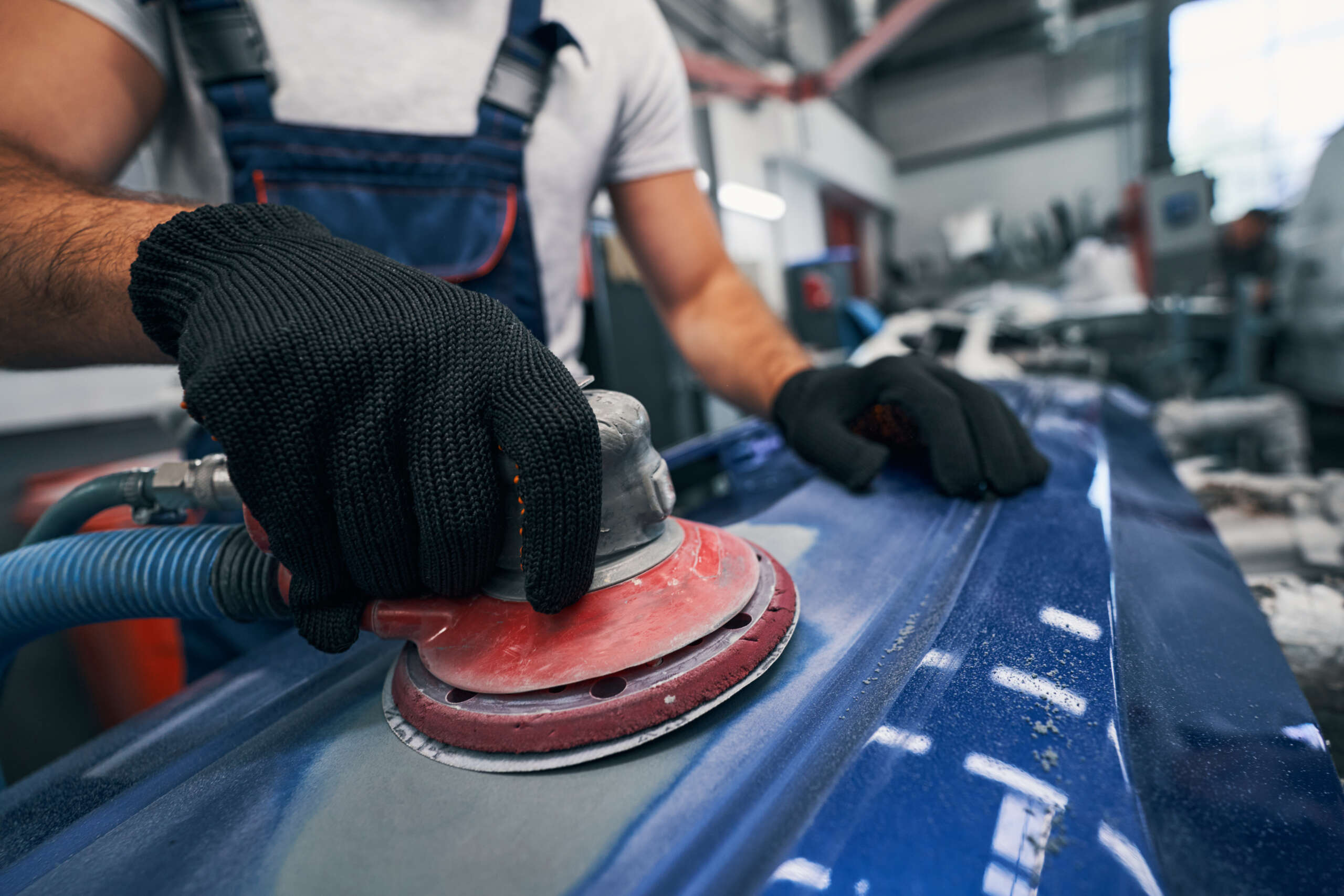 Automobile mechanic in black work gloves using grinder on blue metal surface of car spare part